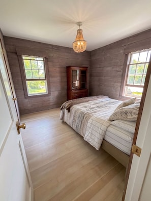 A bedroom with a wooden floor, a bed with patterned bedding, a wooden cabinet, and a chandelier.