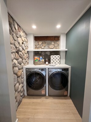 A laundry room with two front-loading washing machines, a shelf with laundry supplies, and a stone wall. - Shelter Island remodeled 1937 Cape Cod Waterfront with Deepwater Dock (Shelter Island)