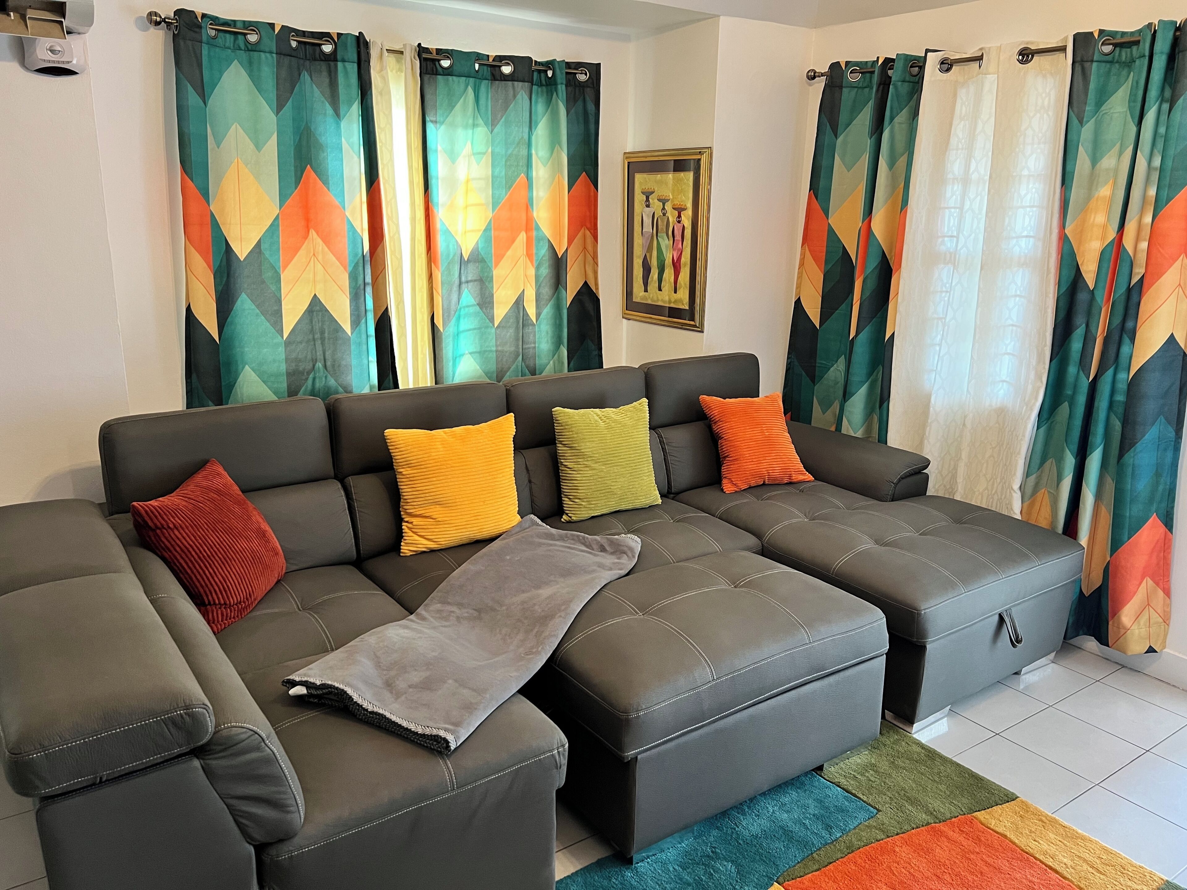 A sectional sofa with multiple cushions, a grey ottoman, and a colorful rug.
