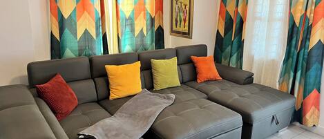 A sectional sofa with multiple cushions, a grey ottoman, and a colorful rug.