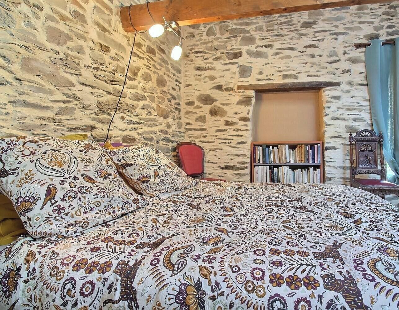 A bedroom with a stone wall, a bed with patterned bedding, a bookshelf, and a chair.