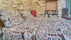 Ein Schlafzimmer mit einer Steinsäule, einem Bett mit gemusterten Textilien, einem Bücherregal und einem Sessel.