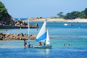 Une scène côti ère avec un voilier, une côte rocheuse, et des personnes qui nagent et se promènent le long de la plage.