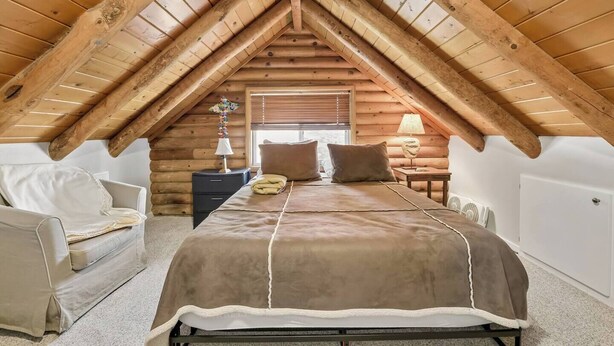 Ein gemütliches Schlafzimmer im Dachgeschoss mit schrägen Wänden, einem Bett mit brauner Decke, einem Sessel, einem Nachttisch und einer Lampe.