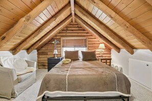 Een knus slaapkamer op zolder met een schuine kap, een bed met een bruine deken, een stoel, een nachtkastje en een lamp.
