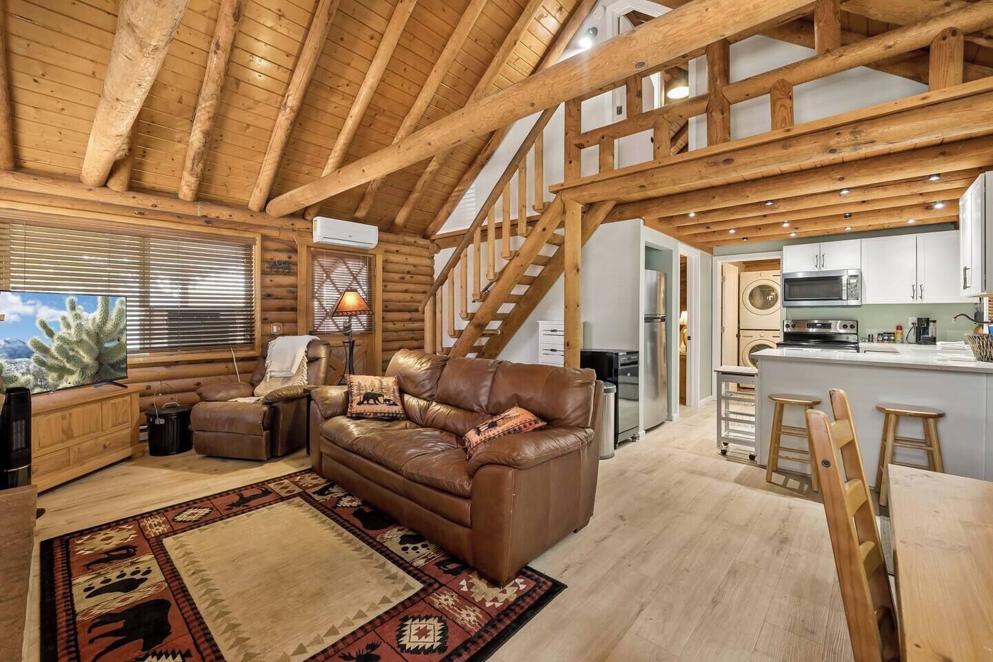 A log cabin interior with a leather sofa, wooden staircase, kitchen area, and a large window with blinds.