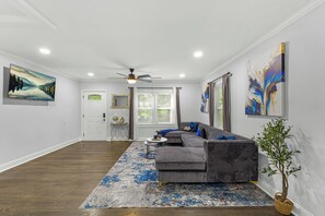 Un salon moderne avec un canapé d’angle gris, un tapis bleu et blanc, un ventilateur de plafond et des œuvres d’art accrochées aux murs.