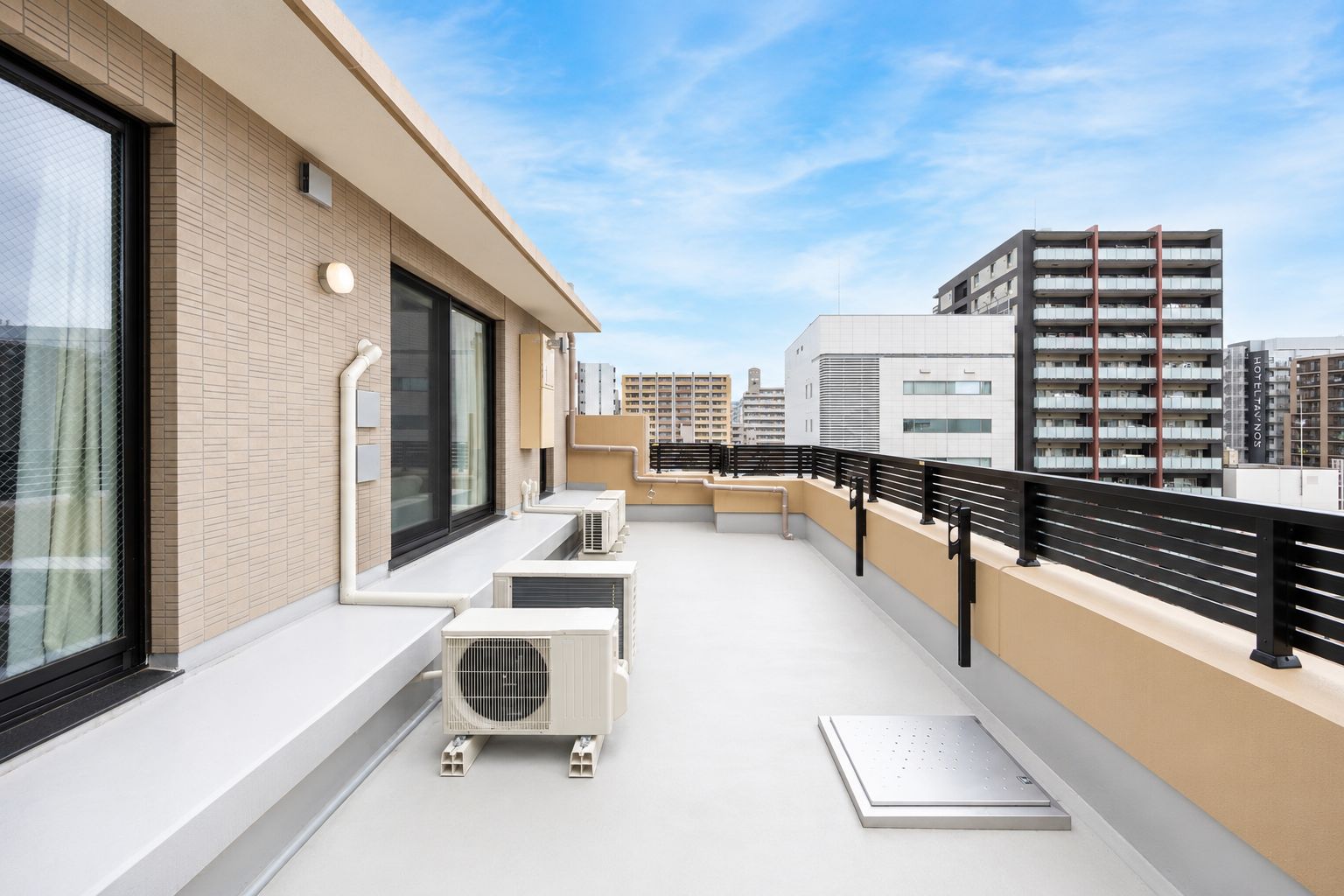 A rooftop terrace with air conditioning units, a black railing, and a view of surrounding buildings.