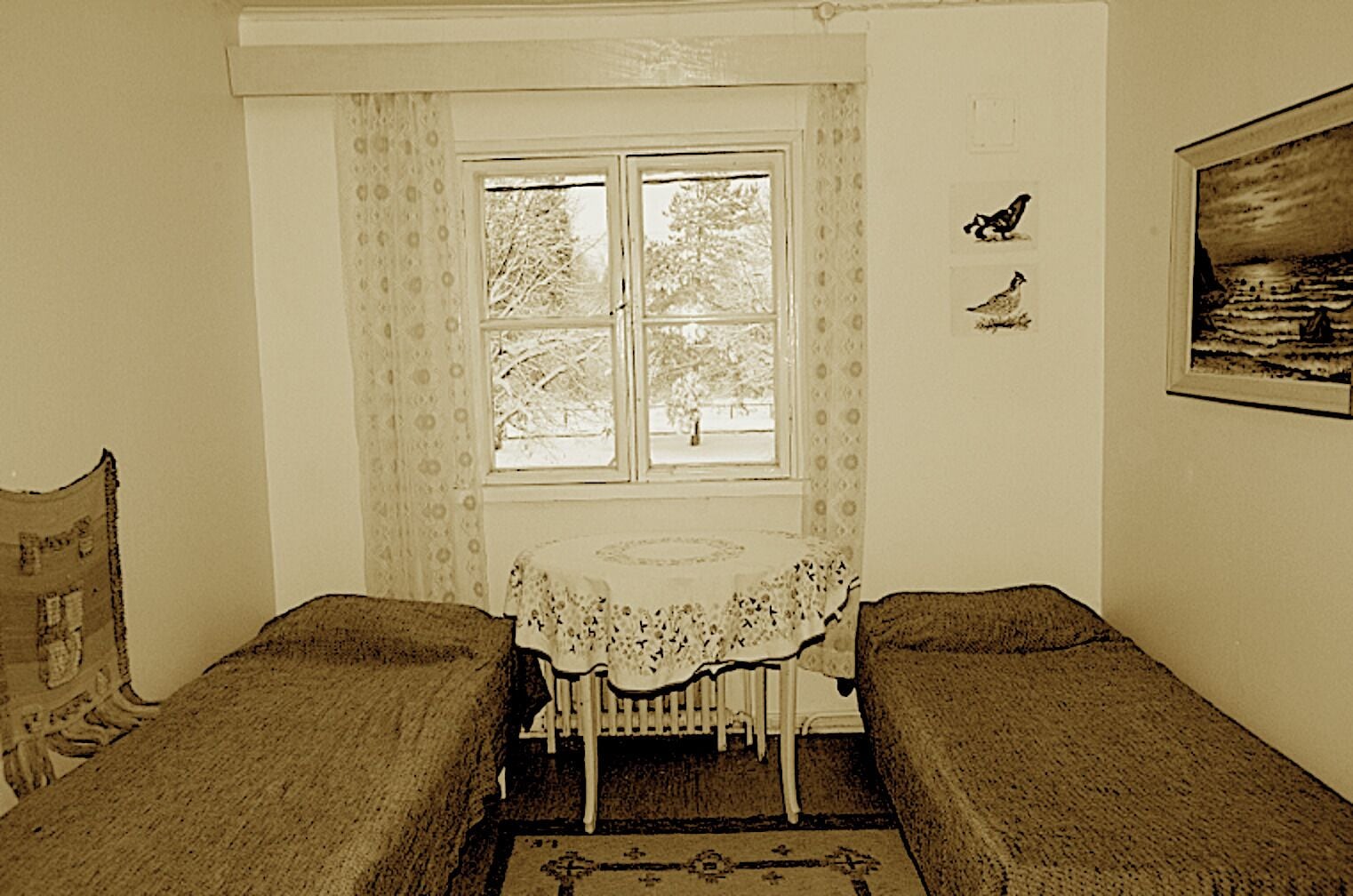 A room with two single beds, a small table with a lace tablecloth, a window with curtains, and a framed picture on the wall.