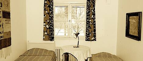 Ein Zimmer mit zwei Einzelbetten, einem Tisch mit Lampe, einem Heizkörper und einem Fenster mit gemusterten Vorhängen.