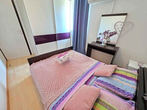 Ein ordentlich bezogenes Bett mit einer rosafarbenen Decke und Kissen, ein Nachttisch mit einer Vase und ein Spiegel an der Wand.