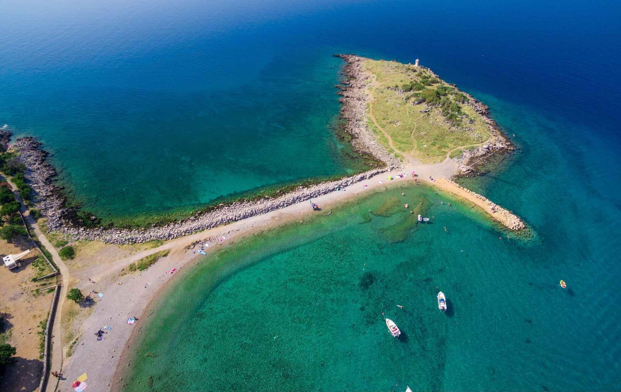 Vue aérienne d’une zone côtière avec une plage, des eaux cristallines et une petite île surmontée d’un phare.