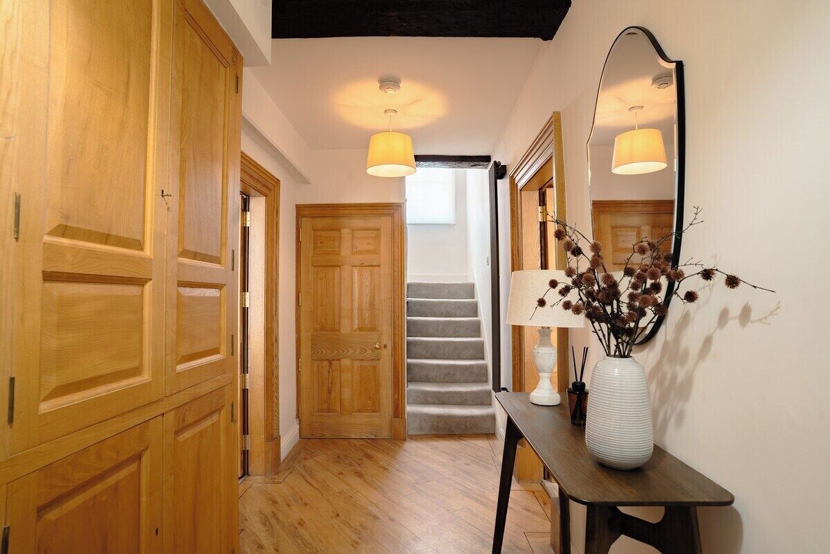 Un couloir avec des portes en bois, un escalier, une table avec un vase et un miroir.