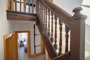 A wooden staircase with balusters, a landing, and a view into a room with a bed and a lamp.