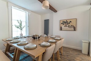 A dining area with a long wooden table set for a meal, a window with a plant, and a painting on the wall.