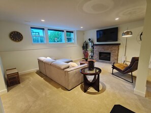 A living room with a sectional sofa, a fireplace, a television mounted above it, and a large window with a view of greenery.