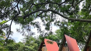 Two wooden A-frame structures with a red roof, set in a grassy area with trees in the background. - Zona Camping El Amanecer (Provincia de Guanacaste)