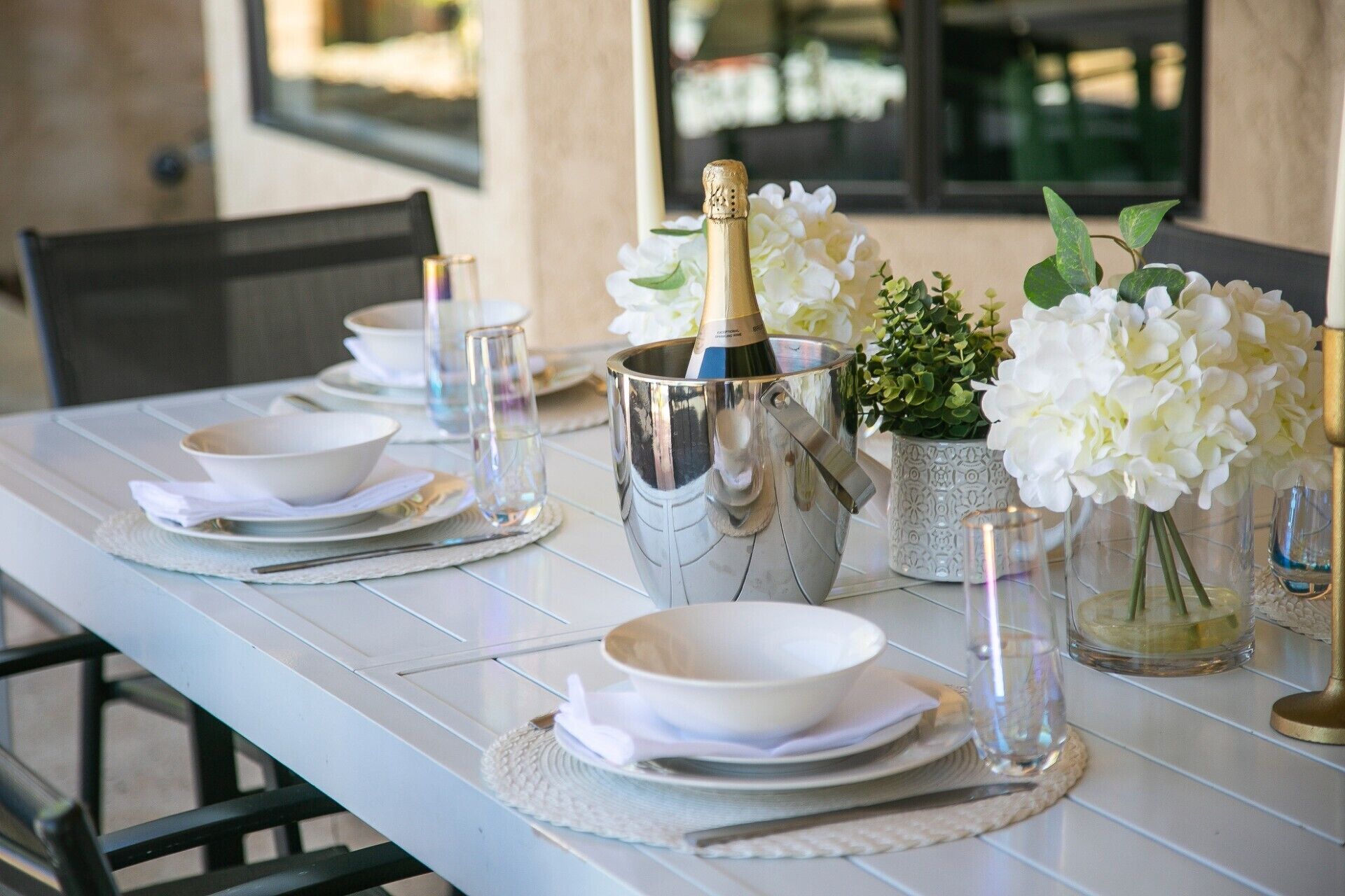 A dining table set with white plates, bowls, and cutlery, a champagne bottle in a silver bucket, and white flowers in a vase.