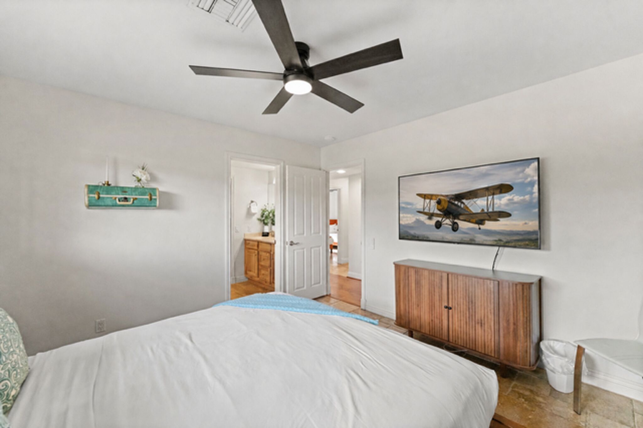 A bedroom with a bed, a ceiling fan, a wooden dresser, a wall-mounted airplane picture, and a door leading to another room.