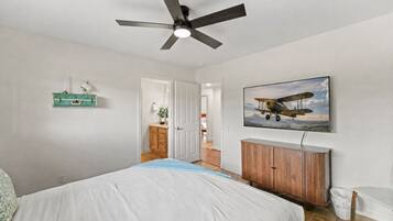 A bedroom with a bed, a ceiling fan, a wooden dresser, a wall-mounted airplane picture, and a door leading to another room.