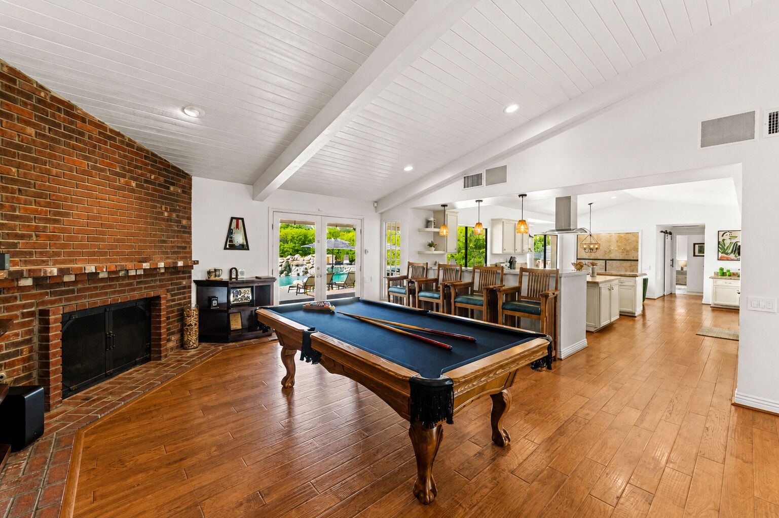 A spacious room with a pool table, a brick fireplace, and a view of a pool and greenery.