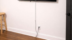 Una habitación con un televisor montado en la pared, un taburete de madera y una cama con colchón blanco.