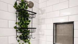Un baño con paredes de azulejos blancos, una ducha con cabezal de mano, una ventana con marco negro y un portalmacetas de pared con hiedra.