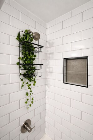 A bathroom with white tiled walls, a shower with a handheld showerhead, a black-framed window, and a wall-mounted plant holder with ivy. - Cozy Home in East Atlanta (Atlanta)
