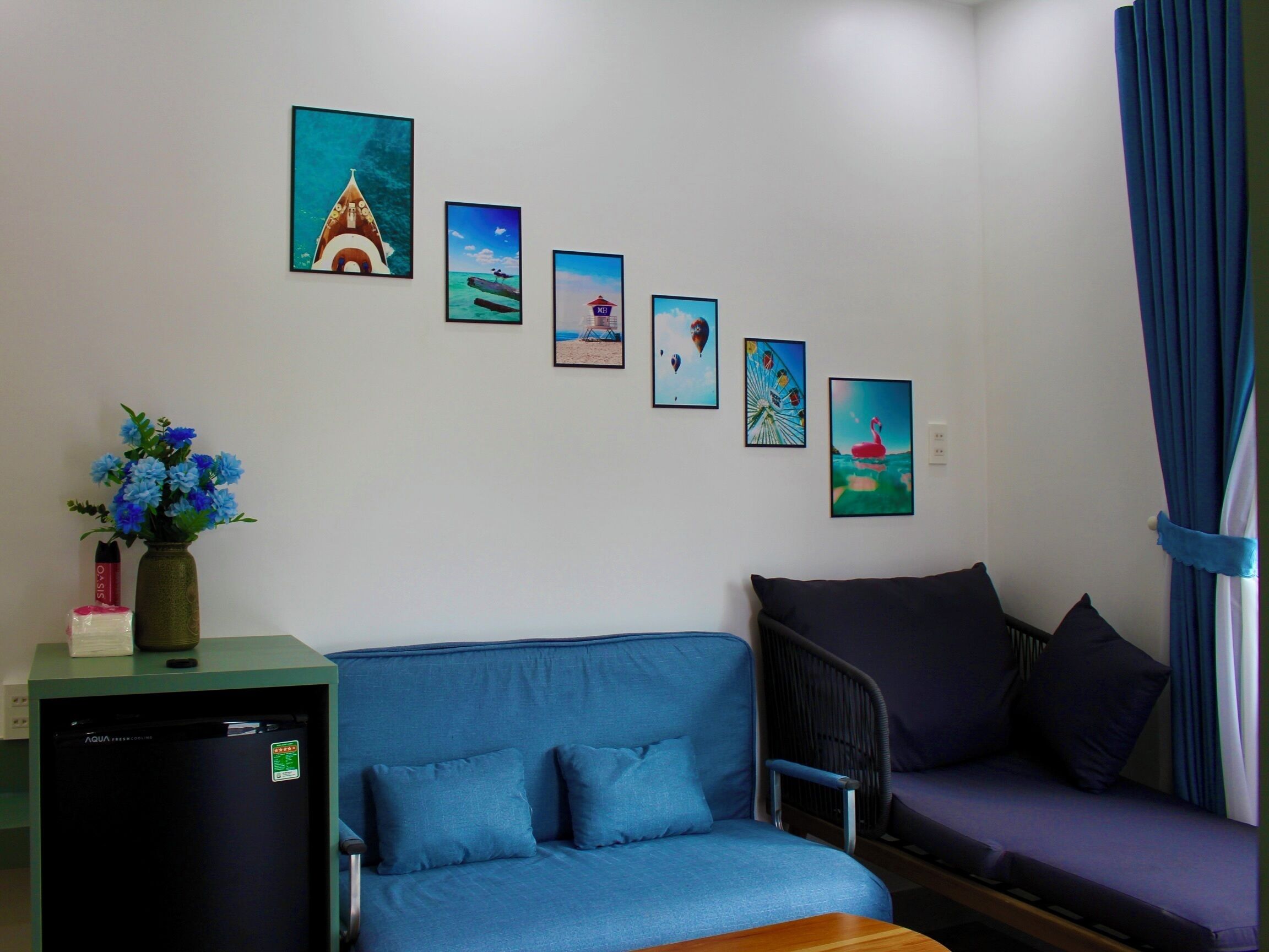 Una habitación con un sofá azul, una mesita con un florero y un refrigerador.