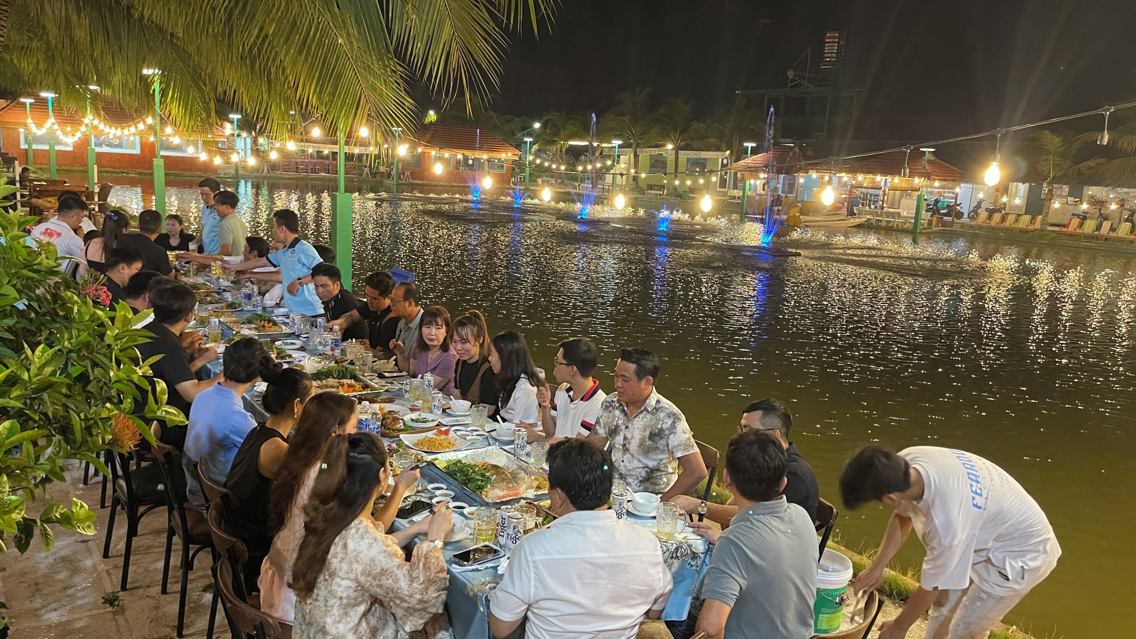Un grupo comiendo junto a un cuerpo de agua, con mesas y sillas, rodeado de follaje y luces colgantes.