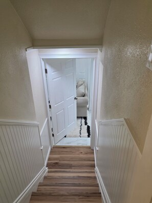 Un couloir doté d’un plancher en bois, de murs blancs et d’une porte donnant sur une pièce où se trouve un tapis noir et blanc.