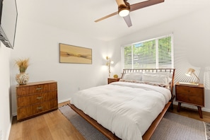 Ein Schlafzimmer mit einem Bett, Holz-Nachttischen, einem Deckenventilator, einem Fenster mit Jalousien, einer Wandleuchte und einer dekorativen Vase.