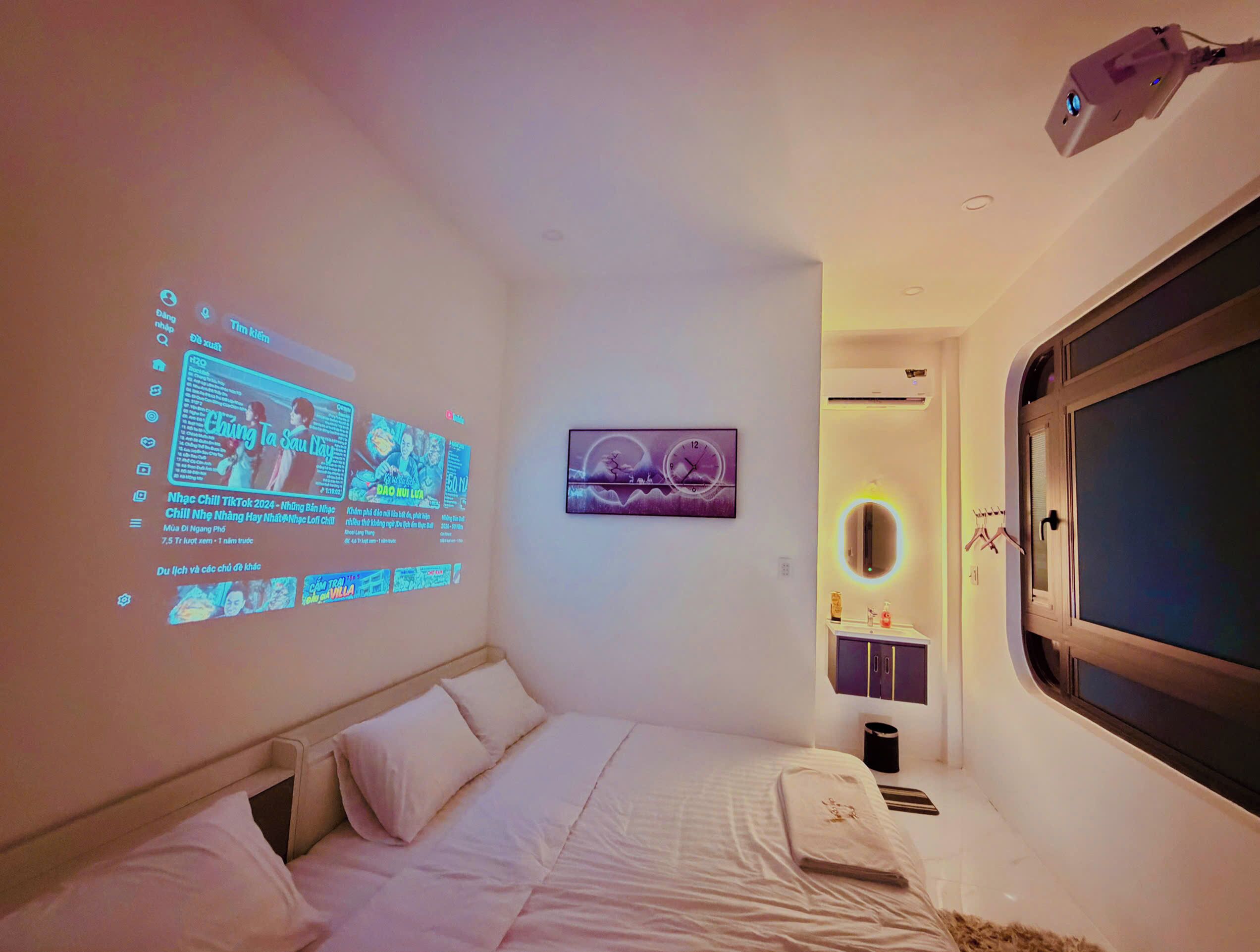A bedroom with a bed, a wall-mounted projector displaying content, a clock, and a window with a view.
