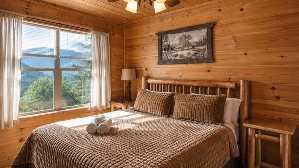 Ein Schlafzimmer einer Holzhütte mit Bett, Nachttisch, Lampe, Deckenventilator und einem Fenster mit Vorhängen.