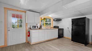 A kitchen with white cabinets, a black refrigerator, a microwave, and a window with a view of trees. - Island Time Bungalow (Steinhatchee)
