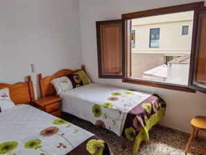 A bedroom with two beds, wooden headboards, a window with a view of buildings, and a small stool. - BERENGUER - Apartment with terrace just 5 minutes from Oliva Beach (Oliva)