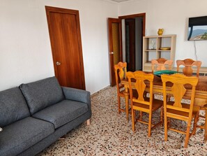 Ein Zimmer mit grauem Sofa, einem Holzespeisetisch mit Stühlen, einem Fernseher an der Wand und einer Tür.