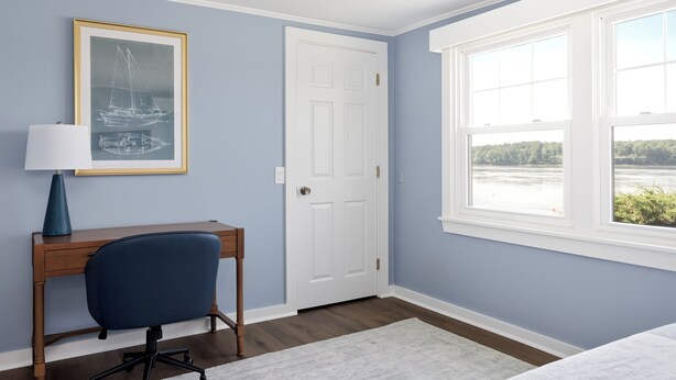 Una habitación con escritorio, silla, cuadro enmarco, una puerta blanca y ventana con vistas.