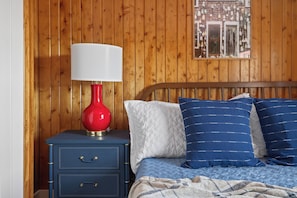 A wooden-paneled room with a bed, a nightstand, a red lamp, and a framed picture on the wall.