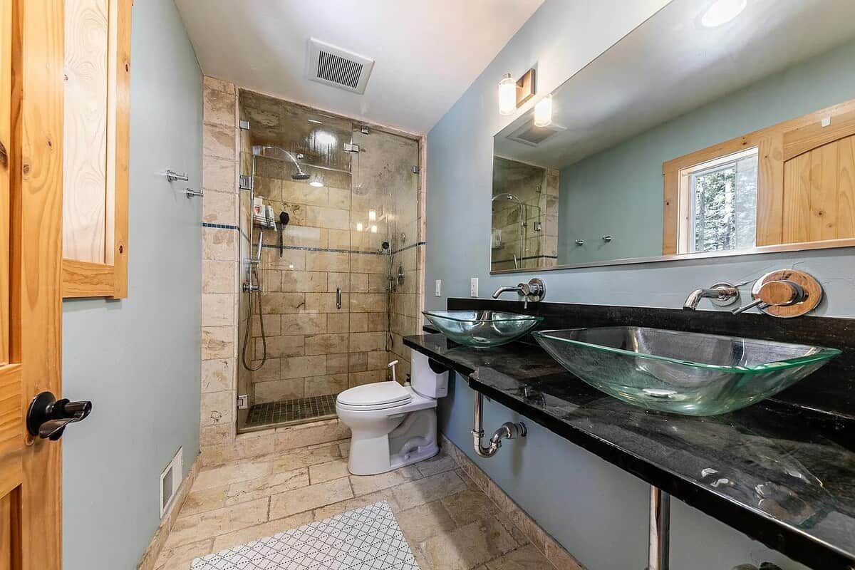 A bathroom with a glass-enclosed shower, a toilet, and a double sink with a glass bowl.