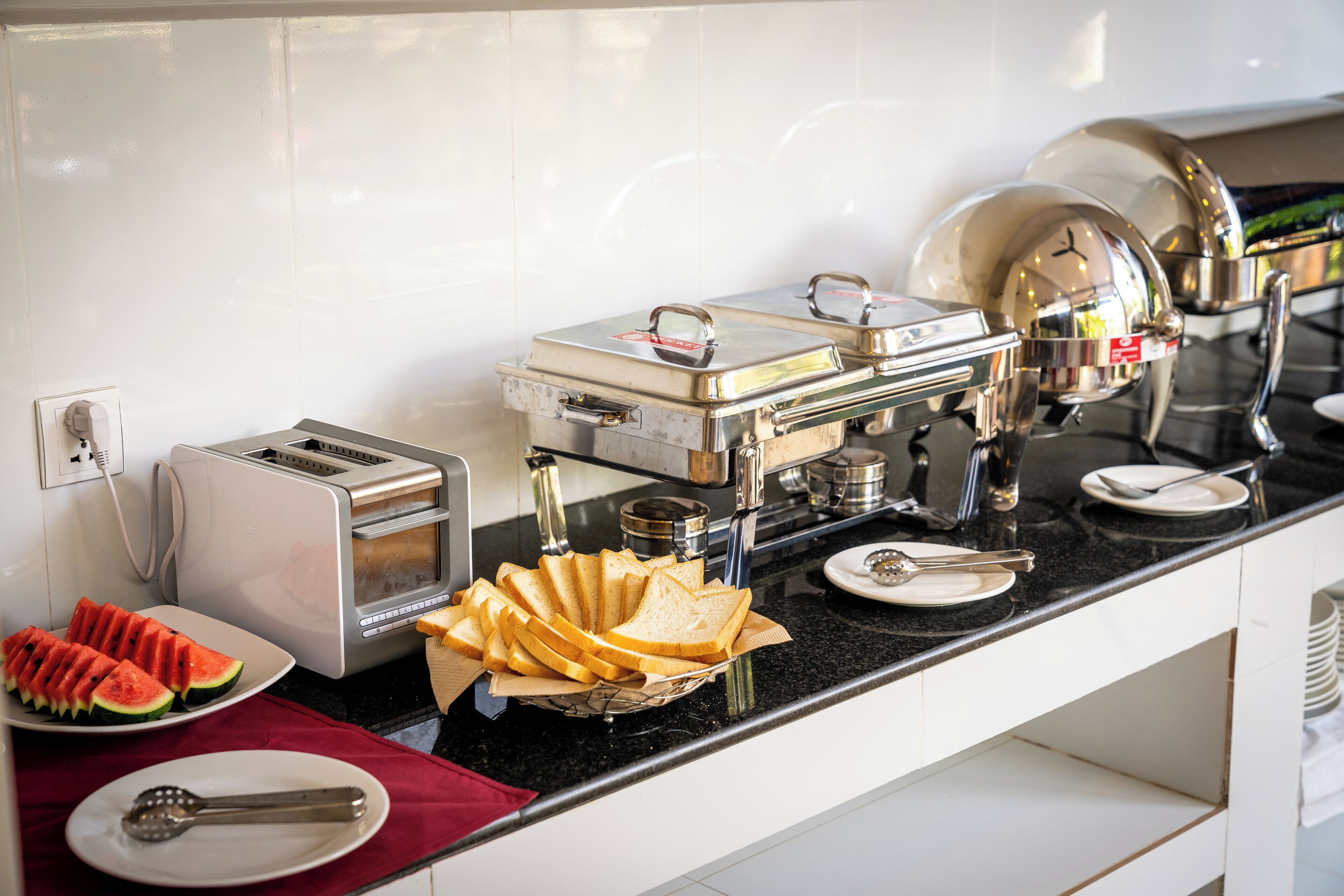 A breakfast buffet setup with a toaster, plates of sliced bread, and a fruit plate.