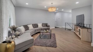 Un salon moderne avec un canapé gris, une table basse, un téléviseur posé sur un meuble en bois et un lustre.