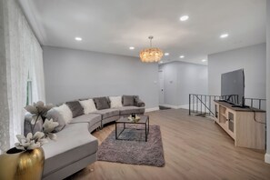 A modern living room with a grey sofa, a coffee table, a TV on a wooden cabinet, and a chandelier. - Luxury 6BR Retreat Near National Harbor & DC (Fort Washington)