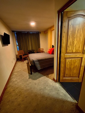 Quarto com cama, porta de madeira, televisão na parede e janela com cortinas.
