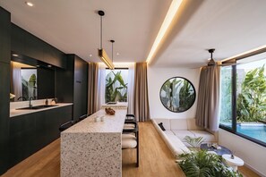 A modern kitchen with a central island, a dining area with a round table, and a seating area with a view of a pool. - Club Suma Villa 5 Bingin (Pecatu)