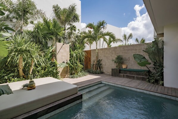 Un área de piscina con cubierta de madera, rodeada de plantas tropicales y un muro de concreto.