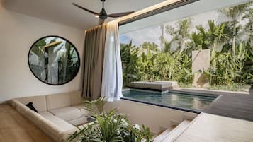 A modern living room with a sofa, a ceiling fan, a round mirror, and a view of a pool and tropical plants.
