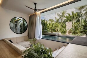 Una sala moderna con un sofá, un ventilador de techo, un espejo redondo y vistas a una piscina y plantas tropicales.