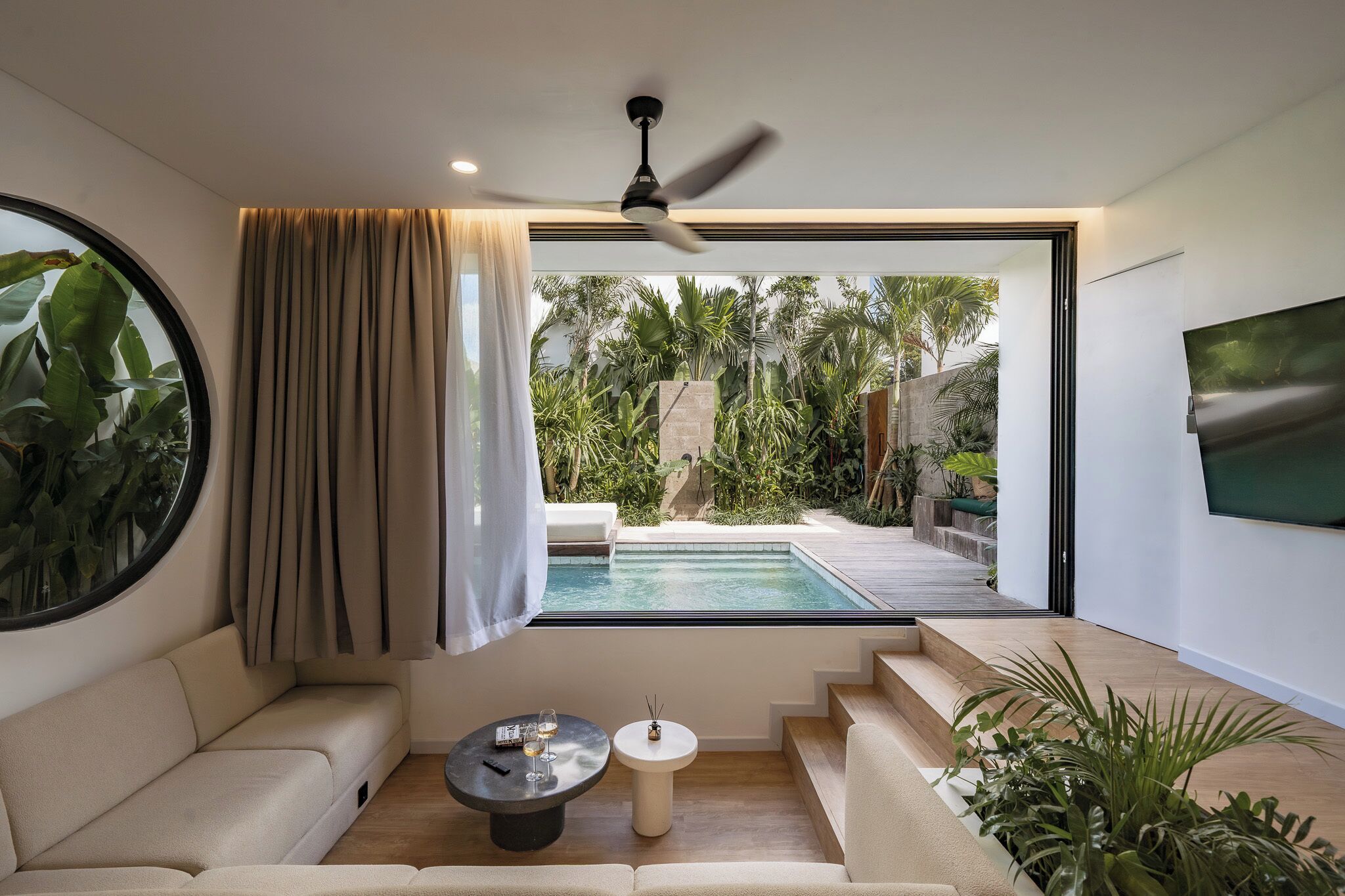 A modern living room with a pool view, a ceiling fan, and a TV mounted on the wall.