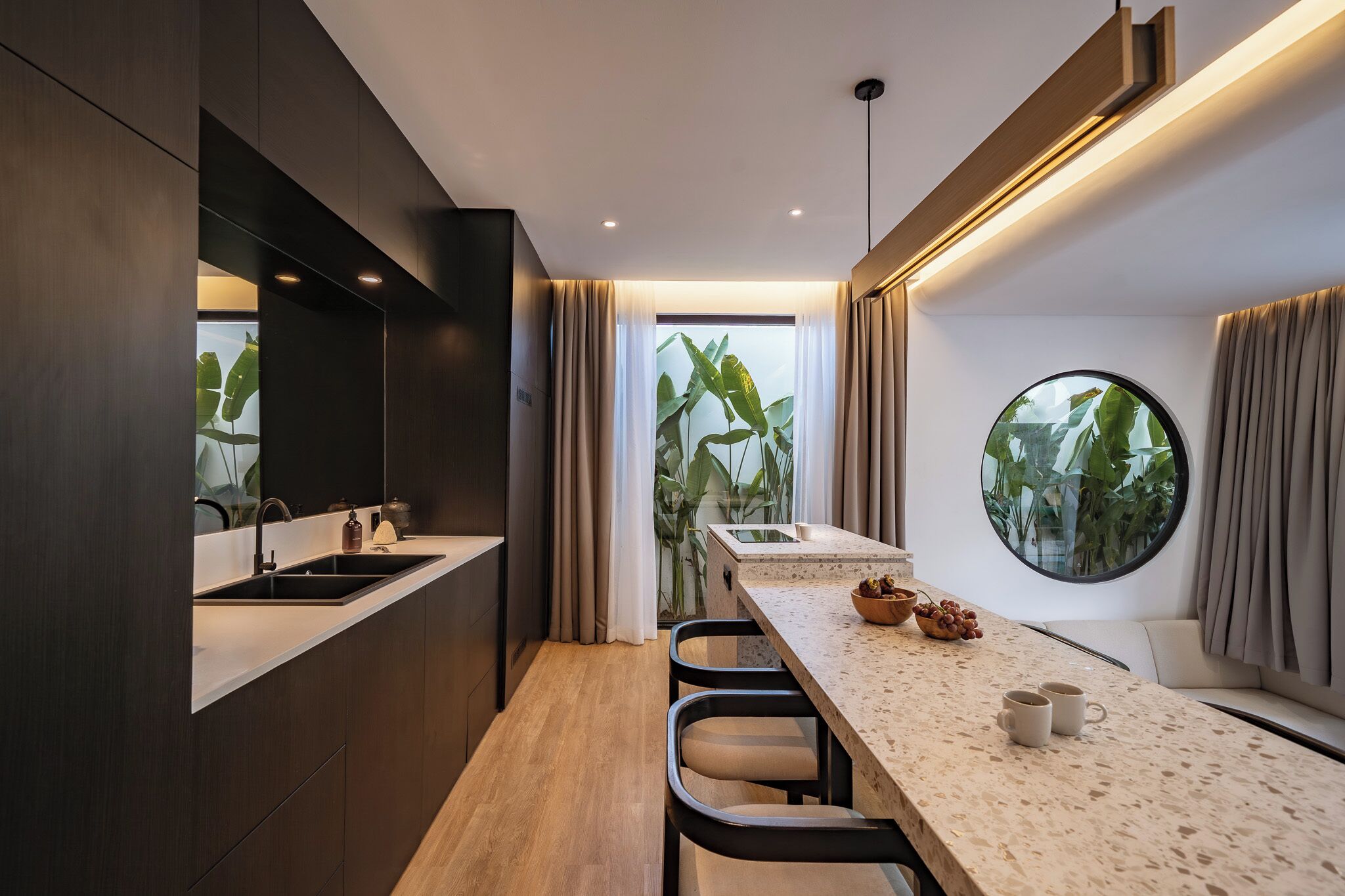 A modern kitchen with a central island, a dining area with a round window, and a view of greenery outside.